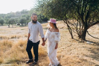 Kırsalda güzel bir çift, beyaz elbiseli yakışıklı sakallı adam ve kadın el ele tutuşuyorlar.