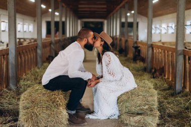 Beyaz, uzun, boho tarzı bir elbise giymiş yakışıklı bir adam ve güzel bir kadın samanlıkta oturup keçi çiftliğinde öpüşüyorlar.