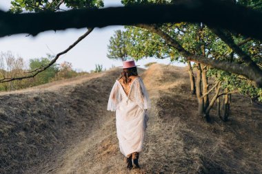 Arkadan görünüşlü, dövmeli, beyaz, uzun bir boho tarzı elbise ve kırsalda pembe şapkalı güzel bir kadın. 