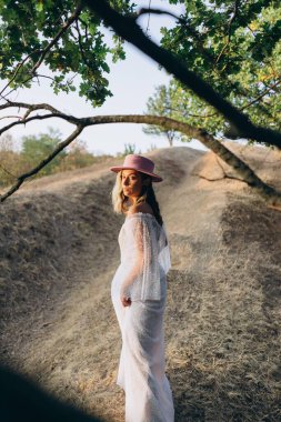 Güzel bir kadın. Renkli saçları ve dövmeli bir vücudu var. Beyaz, boho tarzı bir elbise ve kırsalda pembe bir şapka giyiyor. 