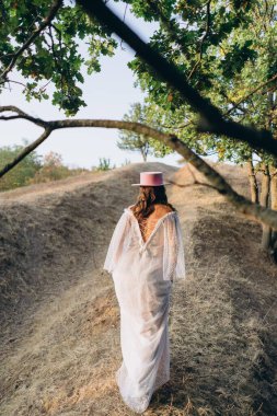 Arkadan görünüşlü, dövmeli, beyaz, uzun bir boho tarzı elbise ve kırsalda pembe şapkalı güzel bir kadın. 