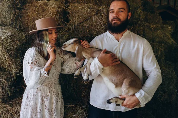 handsome man holding a goat and standing near beautiful woman dressed in a white long boho style dress against of hay