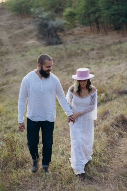 Kırsalda güzel bir çift, beyaz elbiseli yakışıklı sakallı adam ve kadın el ele tutuşuyorlar.