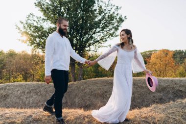 Kırsalda güzel bir çift, beyaz elbiseli yakışıklı sakallı adam ve kadın el ele tutuşuyorlar.