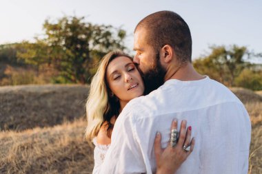 Kırsalda güzel bir çift, yakışıklı sakallı bir adam beyaz elbiseli bir kadına sarılıp onu öpüyor. 