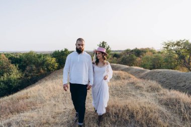 Kırsalda güzel bir çift, beyaz elbiseli yakışıklı sakallı adam ve kadın el ele tutuşuyorlar.