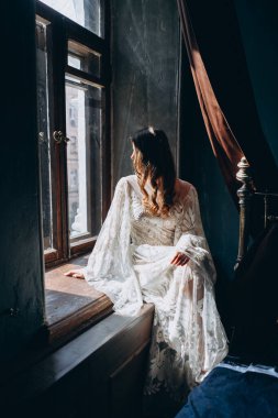 Saçında çiçekler olan, beyaz, bohem bir çiçek desenli elbisesi olan kız yatağın yanındaki pencerede oturuyor ve pencereden dışarı bakıyor.