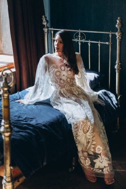 Güzel, beyaz, bohem, şehvetli çiçekli bir elbise giymiş bir kız. Işıklar ve gölgeler içinde yatağında oturuyor ve başka tarafa bakıyor.