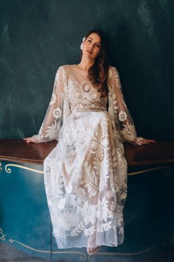 Beyaz, bohem, güzel bir elbise giyen, beyaz saçlı, mavi bir duvarın kenarında oturan ve kameraya bakan bir kız.