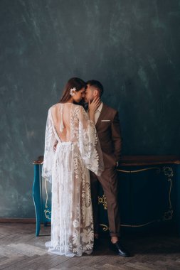 Beyaz bir bohem elbisesi içinde genç ve güzel gelin yarım dönüş yapıp damadını öpüyor. Mavi odada zarif bir takım elbise giymiş.