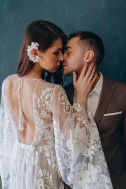 Beyaz bir bohem elbisesi içinde genç ve güzel gelin yarım dönüş yapıp damadını öpüyor. Mavi odada zarif bir takım elbise giymiş.