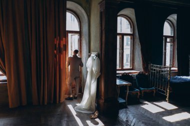 Zarif takım elbiseli bir adam kameraya arkası dönük duruyor ve büyük kemer penceresinden dışarı bakıyor ve adamın yanındaki klasik aynada asılı duran güzel, beyaz, şehvetli tül bir elbise giyiyor.  