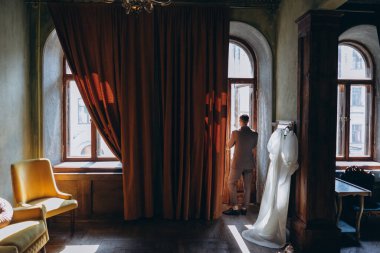 Zarif takım elbiseli bir adam kameraya arkası dönük duruyor ve büyük kemer penceresinden dışarı bakıyor ve adamın yanındaki klasik aynada asılı duran güzel, beyaz, şehvetli tül bir elbise giyiyor.  