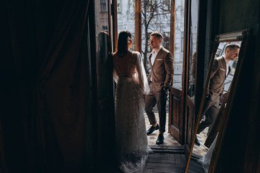 Zarif takım elbiseli bir adam, güzel beyaz, şehvetli bir tül elbise giymiş kız arkadaşının önünde dikiliyor, balkonun kapısında dikiliyorlar ve yansımaları balkonun yanındaki aynada görünüyor.