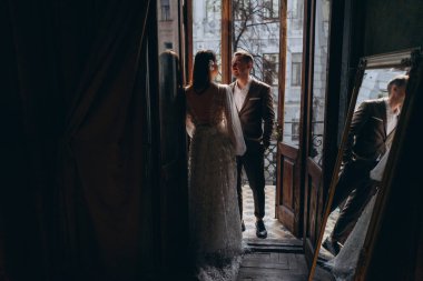 Zarif takım elbiseli bir adam, güzel beyaz, şehvetli bir tül elbise giymiş kız arkadaşının önünde dikiliyor, balkonun kapısında dikiliyorlar ve yansımaları balkonun yanındaki aynada görünüyor.