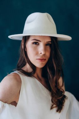 Beyaz elbiseli, beyaz şapkalı bir kız mavi odada kameraya poz veriyor.