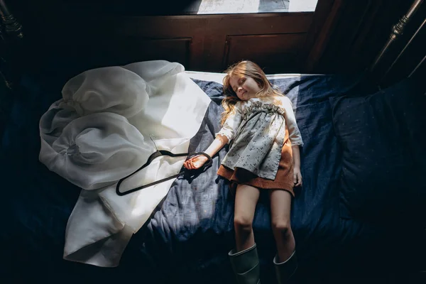 ベージュのヴィンテージのドレスを着た女の子とベッドの上で寝ている緑色のゴム製のブーツ彼女の手の中に黒いハンガーと彼女の近くに横たわっている白い官能的なチュールのドレス — ストック写真