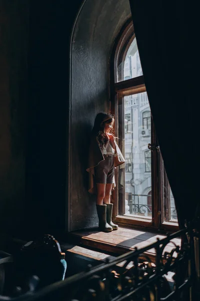 the child in vintage clothes, the little girl in a beige riding hood, shorts, and green rubber boots standing and looking at the big arch window with curtains