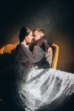 Zarif takım elbiseli bir adam sandalyede oturuyor ve beyaz, güzel, şehvetli bir elbise giymiş bir kız onu pencerenin kenarında sıcak bir ışıkla öpüyor.