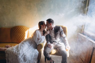 Zarif takım elbiseli adam sandalyede oturuyor ve beyaz, güzel, şehvetli elbisesi olan bir kız dumanlı odanın penceresinde onu öpüyor.