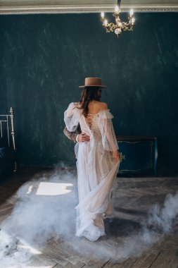 Zarif takım elbiseli bir adam dumanlı odada güzel beyaz tül elbiseli ve şapkalı bir kıza sarılıyor.