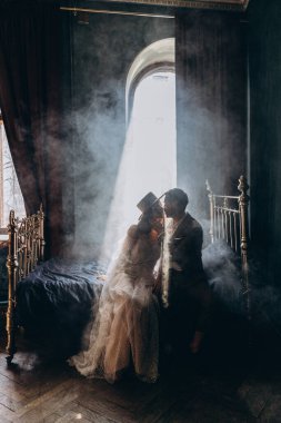 Zarif takım elbiseli bir adam ve güzel, beyaz, şehvetli bir elbise ve şapka giyen bir kız. Yatakta oturmuş, dumanlı odada öpüşüyor.
