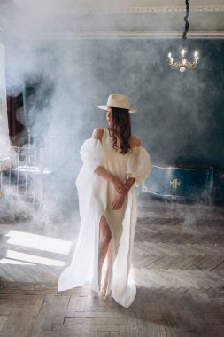  Beyaz, şehvetli elbisesi ve beyaz şapkası olan kız dumanlı odada kameraya poz veriyor.