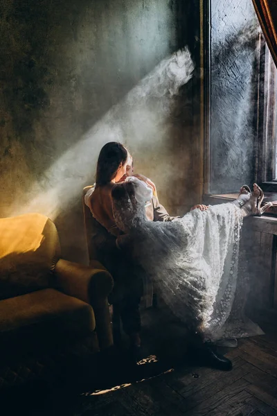 椅子に座っているエレガントなスーツの男と美しい白い官能的なチュールのドレスの女の子暖かい光の中で窓のそばで彼にキス — ストック写真
