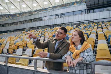 Genç bir çift futbol stadyumunun tribünlerinde dikiliyor ve adam kız arkadaşıyla konuşuyor ve ona bir şey gösteriyor.