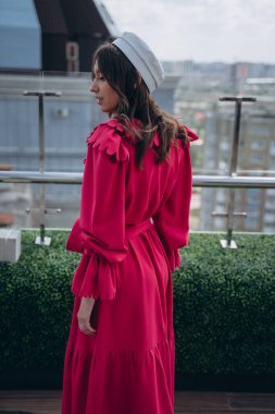 Tasarımcı elbisesi giymiş, parlak yakalı ve beyaz şapkalı, terasta poz veren göz alıcı bir kadının moda fotoğrafı.