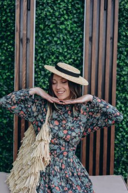 Stüdyodaki beyaz çantayla poz veren şık çiçekli bir elbise ve hasır şapka giymiş güzel bir kadının fotoğrafı.