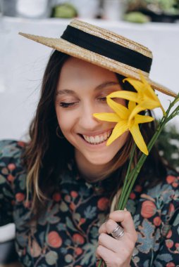 Stüdyoda sarı çiçeklerle poz veren şık çiçekli bir elbise ve hasır şapka giymiş güzel bir kadının fotoğrafı.