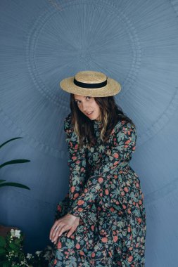 Mavi oda stüdyosunda şık bir çiçekli elbise ve hasır şapka giymiş güzel bir kadının fotoğrafı.