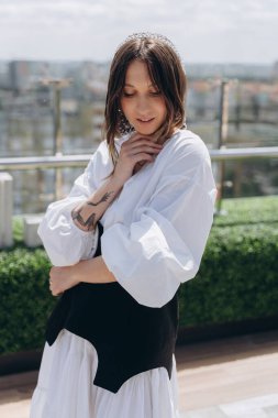 Havalı beyaz bir elbise içinde, açık terasta siyah bir kemerle poz veren çekici bir kadının moda fotoğrafı.