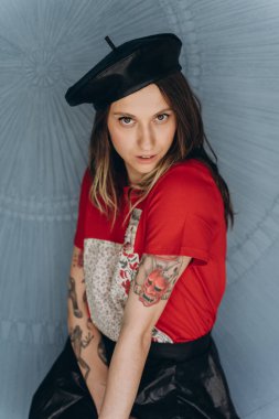 Kollarında dövmeleri olan, kırmızı bluz ve siyah etek giymiş, mavi arka planda poz veren arsız kız manzarası.