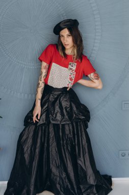 Kollarında dövmeleri olan, kırmızı bluz ve siyah etek giymiş, mavi arka planda poz veren arsız kız manzarası.