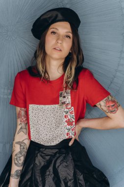 Kollarında dövmeleri olan, kırmızı bluz ve siyah etek giymiş, mavi arka planda poz veren arsız kız manzarası.