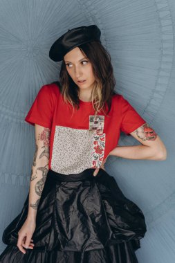 Kollarında dövmeleri olan, kırmızı bluz ve siyah etek giymiş, mavi arka planda poz veren arsız kız manzarası.