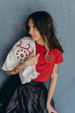 Kollarında dövmeleri olan, kırmızı bluz ve siyah etek giymiş, mavi arka planda poz veren arsız bir kız.