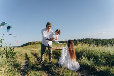 Happy young family spending  time together in nature, beautiful woman in white guipure dress with husband and their daughter walking  in the field