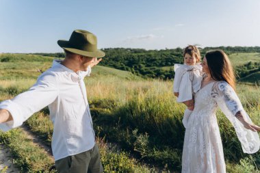 Happy young family spending  time together in nature, beautiful woman in white guipure dress with husband and their daughter walking  in the field