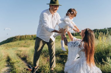 Happy young family spending  time together in nature, beautiful woman in white guipure dress with husband and their daughter walking  in the field