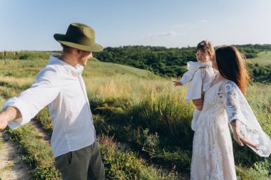 Happy young family spending  time together in nature, beautiful woman in white guipure dress with husband and their daughter walking  in the field