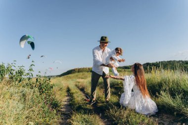 Happy young family spending  time together in nature, beautiful woman in white guipure dress with husband and their daughter walking  in the field