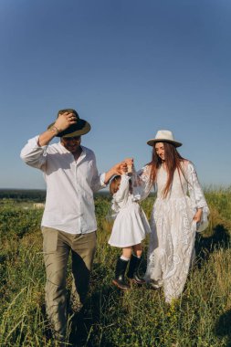 Happy young family spending  time together in nature, beautiful woman in white guipure dress with husband and their daughter walking  in the field