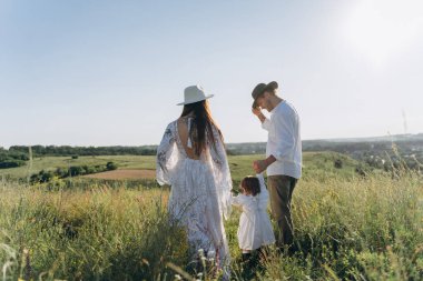Happy young family spending  time together in nature, beautiful woman in white guipure dress with husband and their daughter walking  in the field