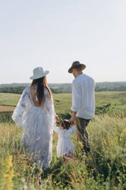 Happy young family spending  time together in nature, beautiful woman in white guipure dress with husband and their daughter walking  in the field
