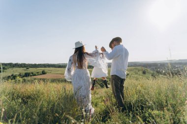 Happy young family spending  time together in nature, beautiful woman in white guipure dress with husband and their daughter walking  in the field