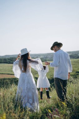 Happy young family spending  time together in nature, beautiful woman in white guipure dress with husband and their daughter walking  in the field