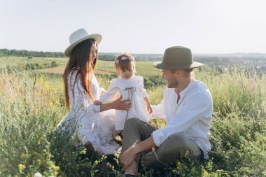 Happy young family spending  time together in nature, beautiful woman in white guipure dress with husband and their daughter sitting in the field